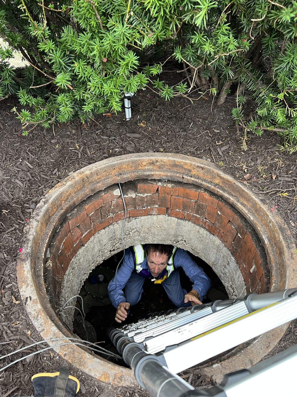 Technician in a safety vest at the bottom of a brick meter vault, wiring a sensor, seen from above