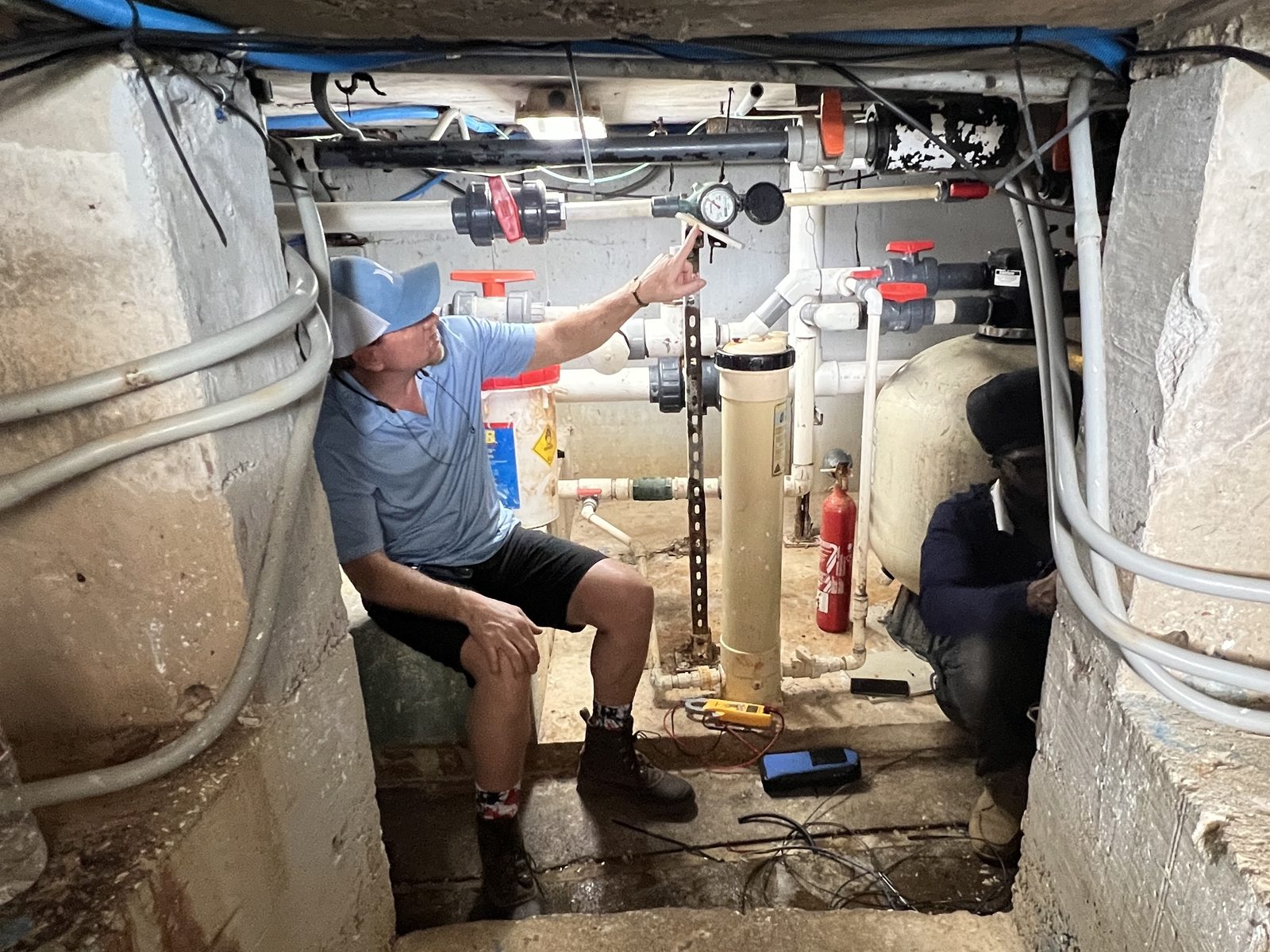 Technician pointing at a flow meter and pressure gauge among pipes in a cramped underground pump room